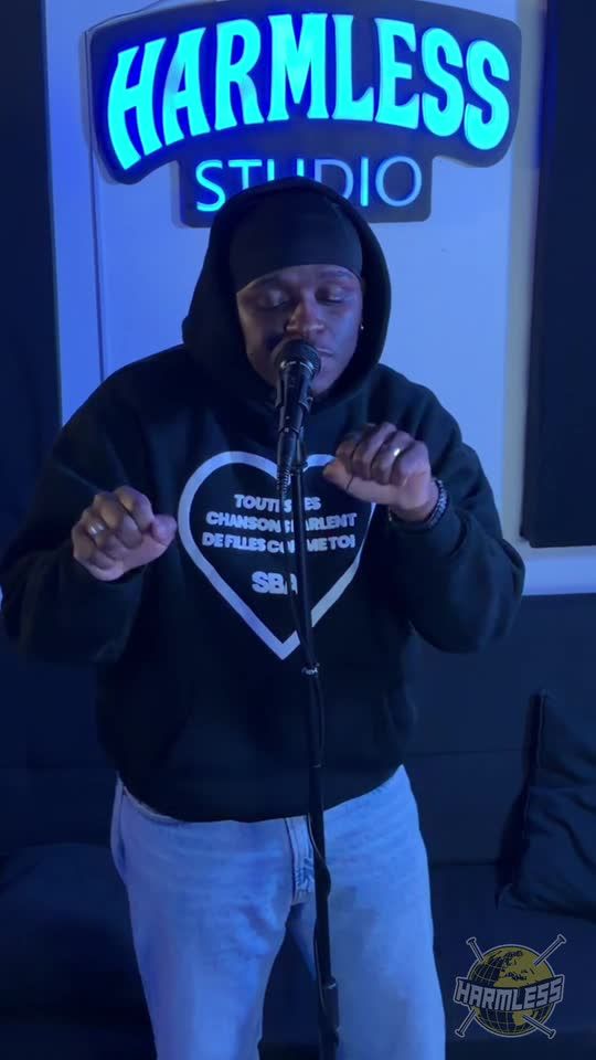 Artist in a black beanie and hoodie performing at the microphone under the glowing Harmless Studio sign