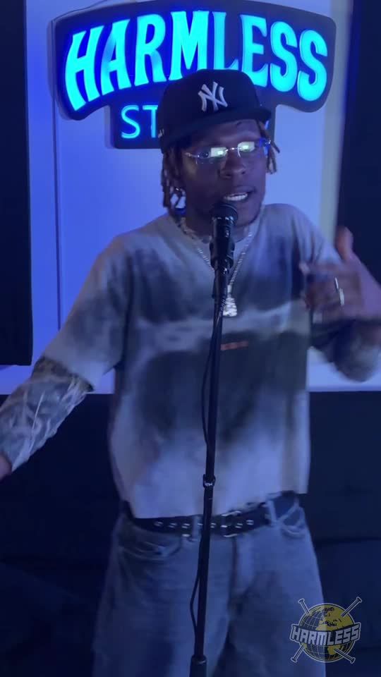 Rapper in a New York cap and tinted glasses performing at the microphone under the Harmless Studio sign