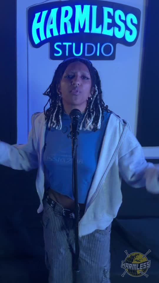 Artist with blonde-tipped braids performing at the microphone under the Harmless Studio sign