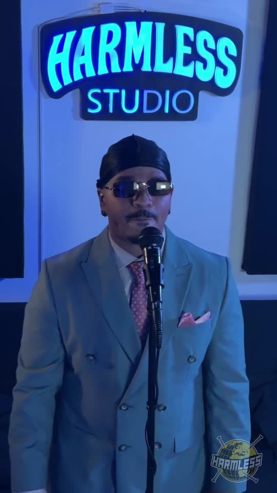 Rapper in a light green suit and pink tie performing at the microphone under the Harmless Studio sign