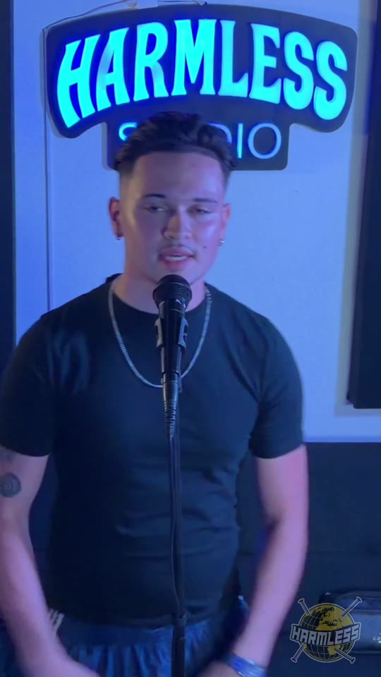 Artist in a black t-shirt performing at the microphone under the Harmless Studio sign